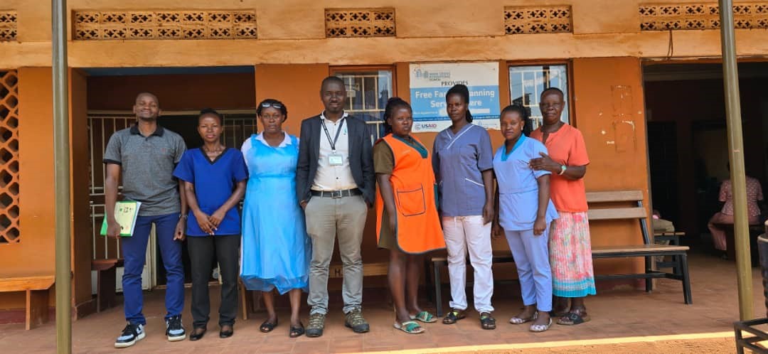 A group photo of the staff of Mutundwe Health Centre II, including the in-charge, Ms. Prossy Naluggwa (in a plastic apron) with Dr. Geoffrey Kisule (middle) and Mr. Allan Ssembuusi (extreme right) after a successful facility visit.