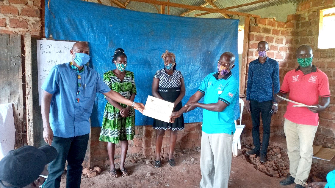 Dr. David Musoke (1st left) handing over a certificate of participation to one of the trained CHWs. Standing behind are health workers and CHWs trainers Doreen Nabwire(L) and Aidah Nakate(R). Henry Bugembe (1st right) and Henry Kajubi (2nd right) are field mobilizers.